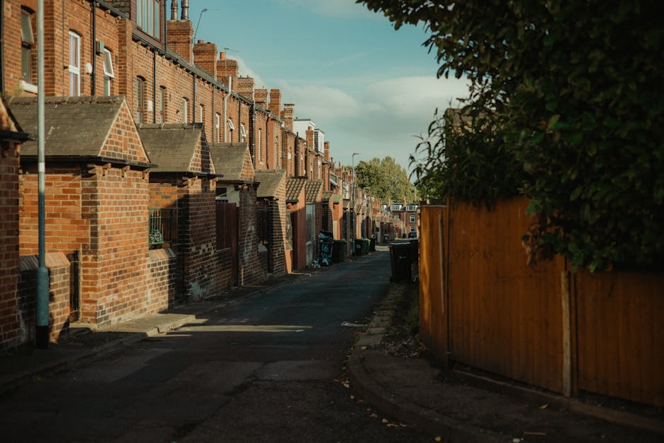 A white household waste collection vehicle is parked on a paved street in front of a row of multi-story buildings with diverse architectural styles, including brick and stucco facades with large windo